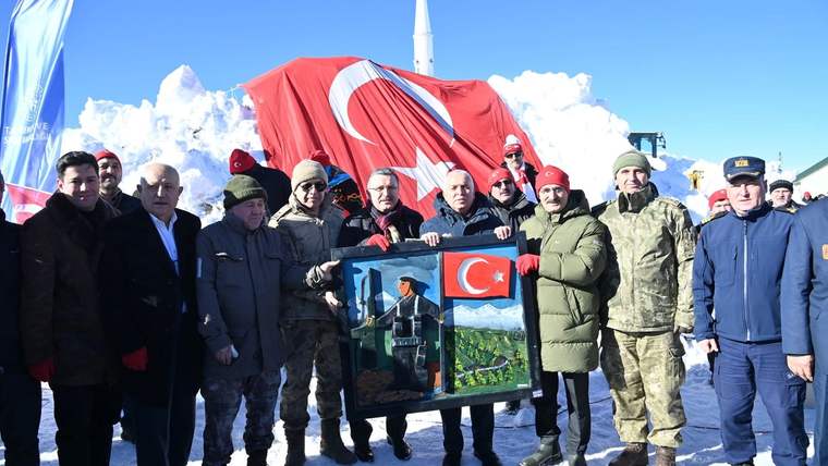 Karlı alanda bir grup insan, Türk bayrağı önünde üzerinde bayrak ve manzara bulunan çerçeveyi tutuyor.