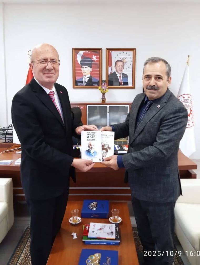 Bir ofiste iki erkek, ellerinde üzerinde “Akif Ersoy” yazılı kitaplarla birlikte duruyor; arkada portreler bulunuyor.