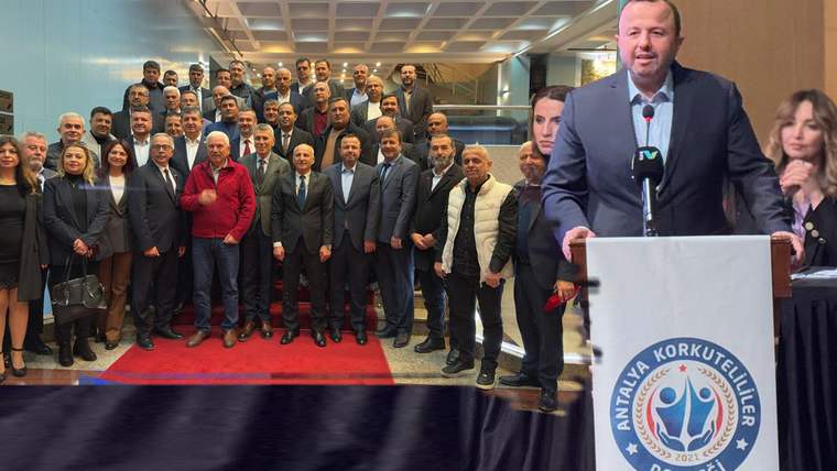 Kalabalık bir grup salonda poz verirken, bir kişi üzerinde Antalya Korkuteliler yazan kürsüde konuşuyor.
