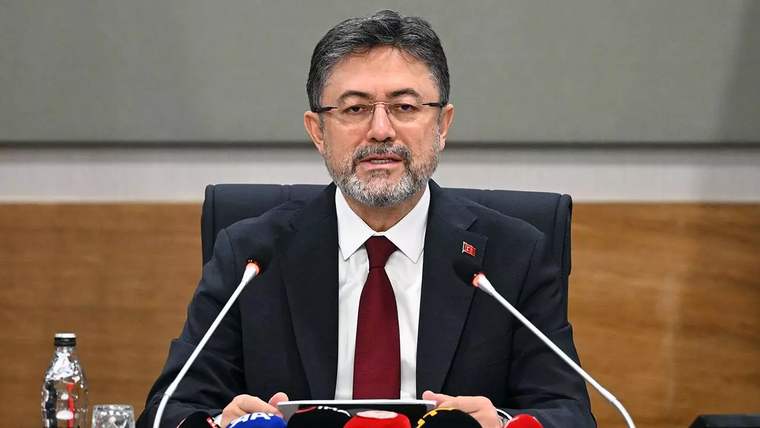 Masa başında oturan takım elbiseli bir kişi, önündeki mikrofon ve basın mikrofonları arasında konuşuyor.