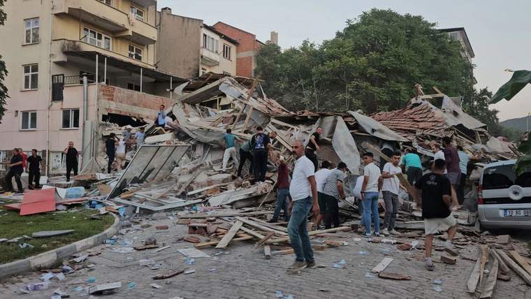 Yıkılmış binaların enkazı önünde çok sayıda kişi molozların arasında duruyor; çevrede sağlam yapılar görülüyor.