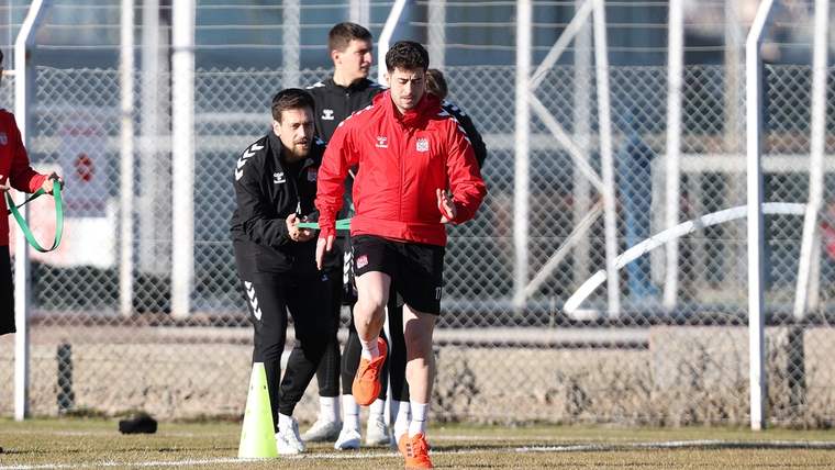 Kırmızı forma giymiş bir futbolcu, antrenman sahasında koşuyor; arka planda diğer oyuncular ve antrenman ekipmanları var.