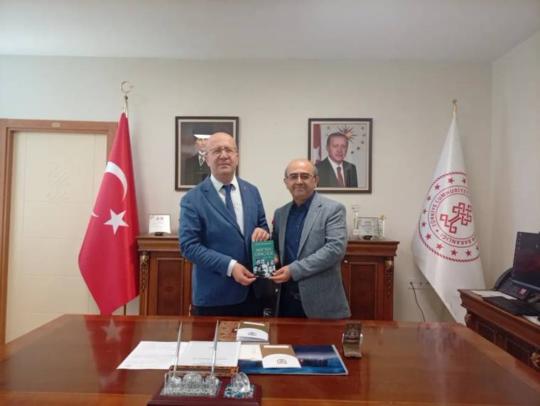 Ofis ortamında iki erkek, Türk bayrağı ve kurumsal flamaların önünde bir kitapçık tutuyor.