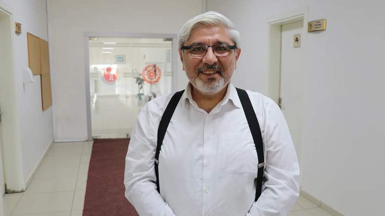 Beyaz gömlek giymiş, gözlük takan bir adam, gülümseyerek poz veriyor.