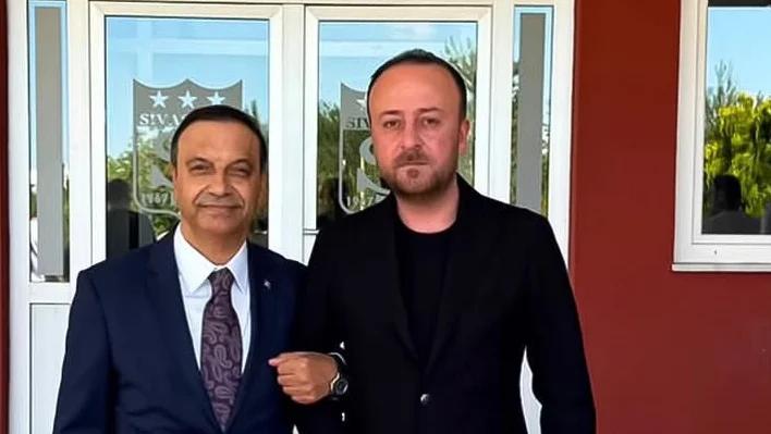 Bir binanın girişinde, cam kapılardaki yıldızlı takım arması önünde duran iki erkek görülüyor.