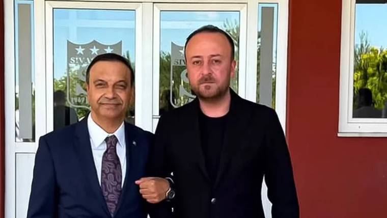 Bir binanın girişinde, cam kapılardaki yıldızlı takım arması önünde duran iki erkek görülüyor.