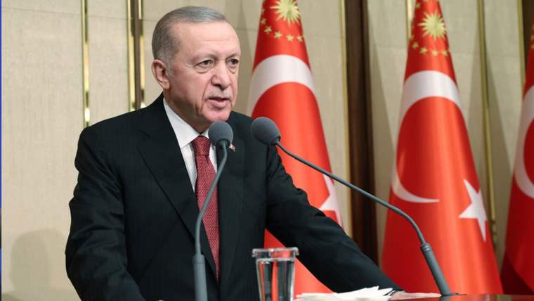 Kürsüde konuşan bir kişi, arkasında ay yıldızlı bayraklar ve önünde mikrofonlar bulunuyor.