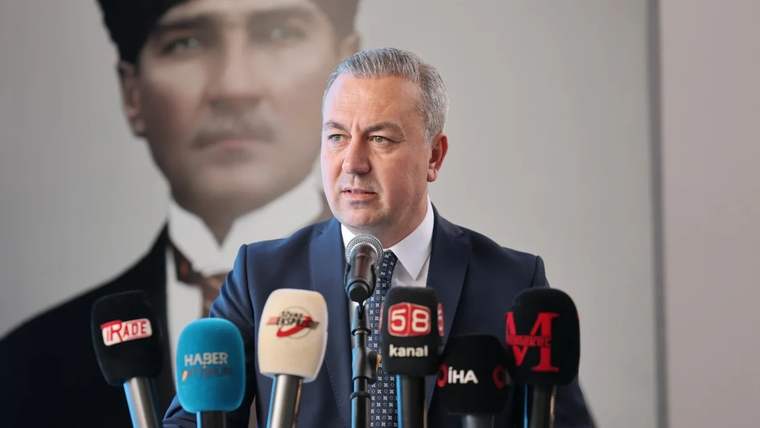 Mikrofonun önünde konuşan bir kişi, arka planda büyük bir portre yer alıyor.