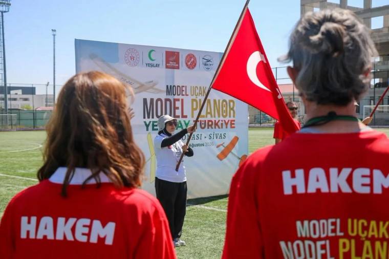 Sahada iki hakem tişörtlü kişi, model uçak ve planör yazılı pankart önünde konuşan kişiyi dinliyor.