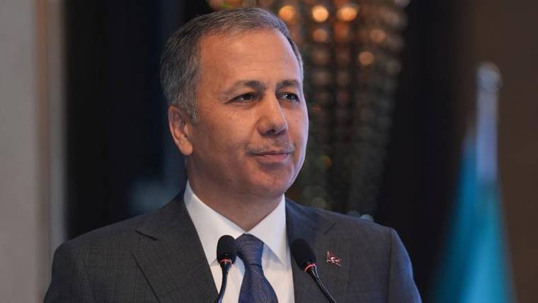 Mikrofonların önünde takım elbiseli bir erkek, yakasında küçük kırmızı rozetle kameraya bakıyor.