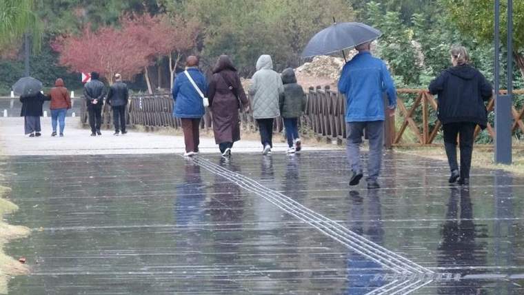 Yağmurlu havada şemsiyelerle yürüyen insanlar, ıslak bir park yolunda ilerliyor.