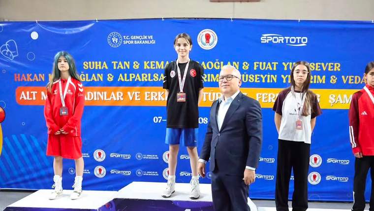 Madalyalı dört genç sporcu podyumda dururken, önlerinde takım elbiseli bir kişi ve arkada mavi pankart görülüyor.