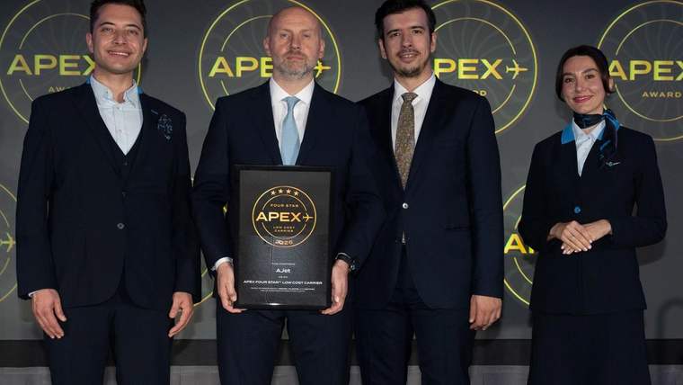 Üç takım elbiseli erkek ve üniformalı bir kadın, APEX ödül panosu önünde; ortadaki erkek plaket tutuyor.