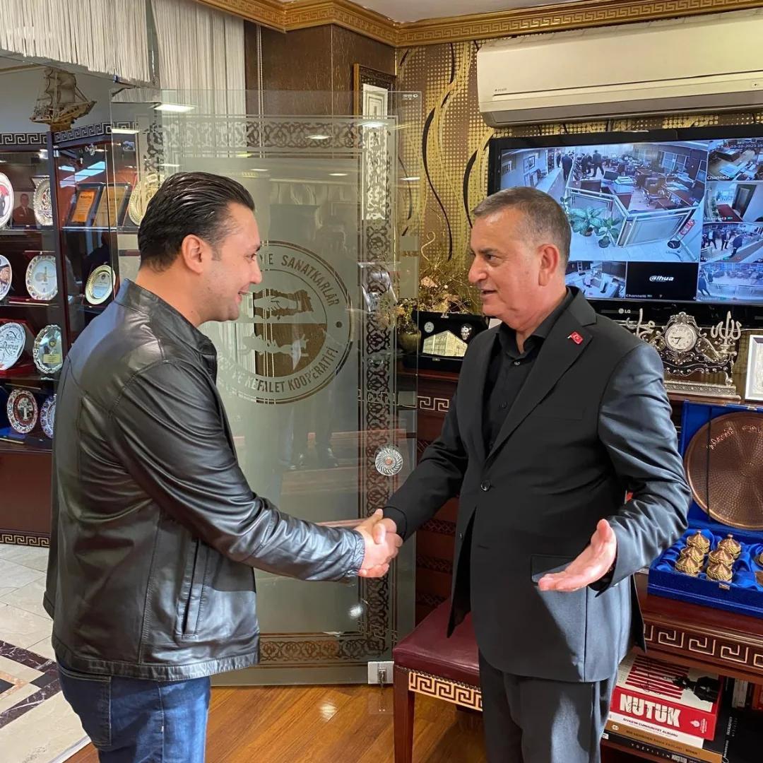 İç mekânda iki erkek tokalaşırken arkalarında ödül vitrinleri ve güvenlik kameralarının görüntülendiği ekran bulunuyor.
