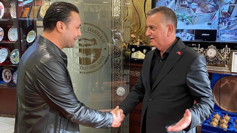 İç mekânda iki erkek tokalaşırken arkalarında ödül vitrinleri ve güvenlik kameralarının görüntülendiği ekran bulunuyor.