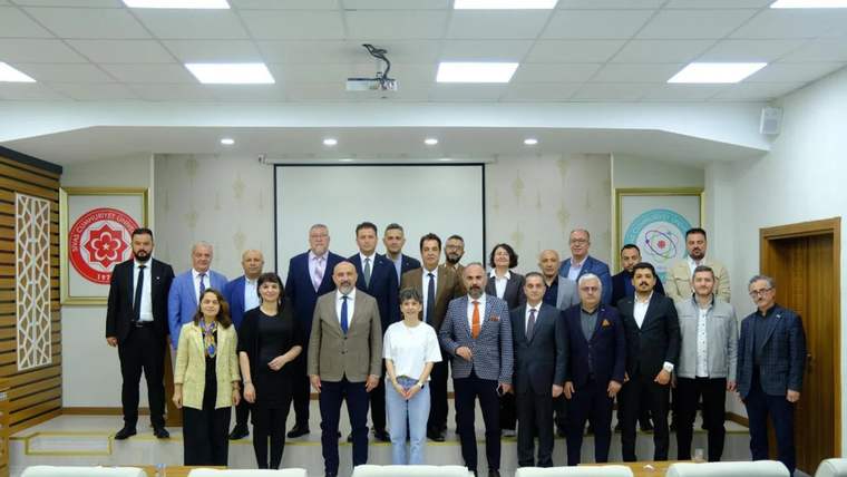 Toplantı salonunda, projeksiyon perdesi önünde farklı yaşlardan yaklaşık 20 kişi birlikte duruyor.