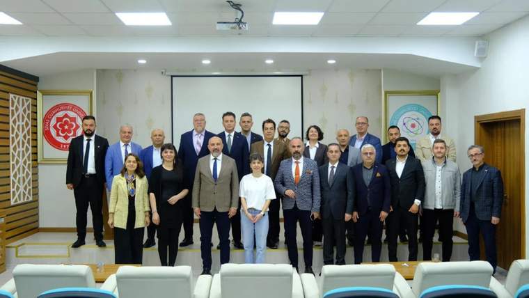 Toplantı salonunda, projeksiyon perdesi önünde farklı yaşlardan yaklaşık 20 kişi birlikte duruyor.