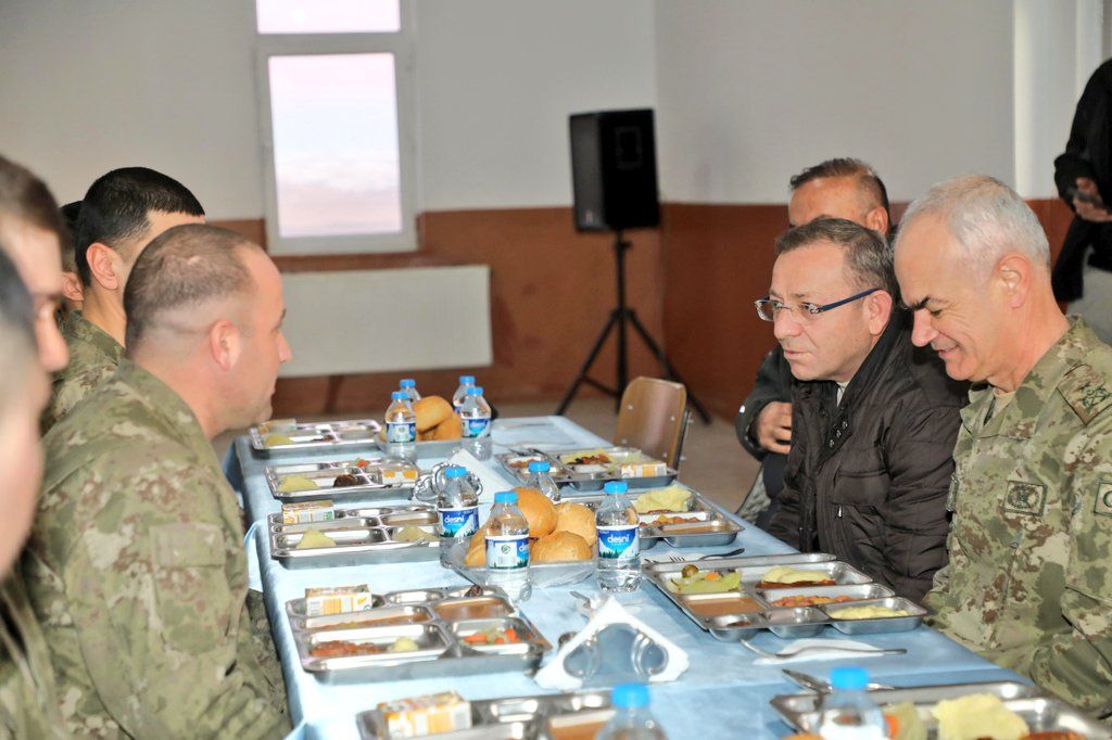 Askeri üniforma giymiş birkaç kişi, iftar sofrasında oturuyor. Masada yemek ve içecekler var.