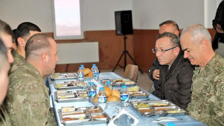 Askeri üniforma giymiş birkaç kişi, iftar sofrasında oturuyor. Masada yemek ve içecekler var.