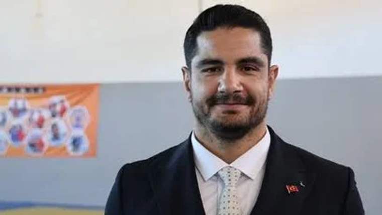 Koyu takım elbise giymiş, gülümseyen bir erkek sporcu, arka planda spor salonu görünmekte.