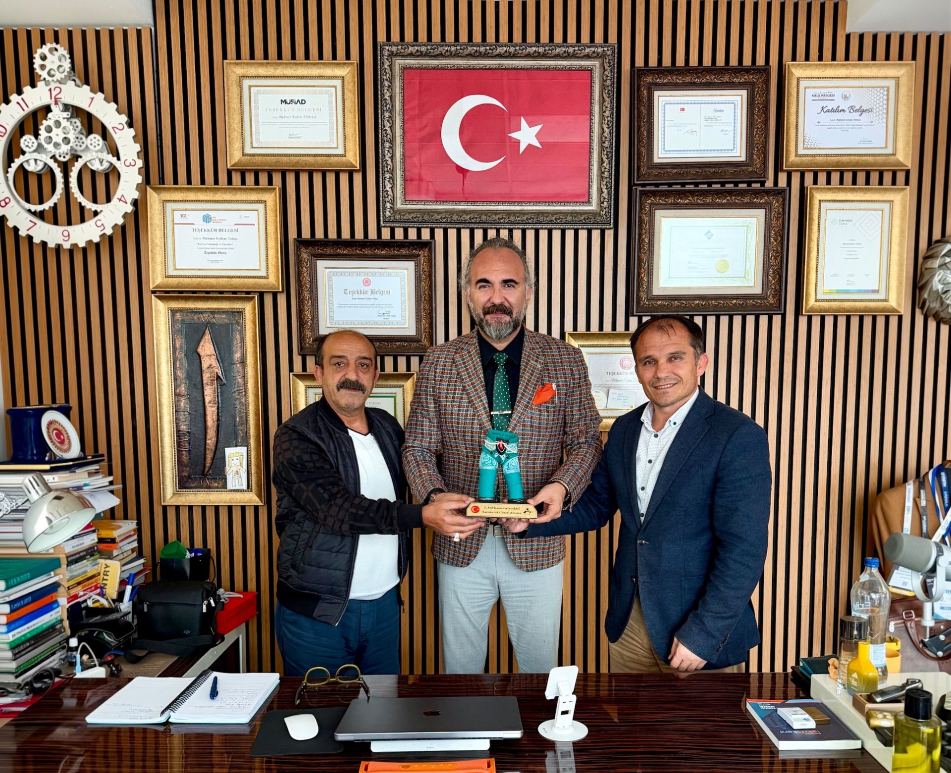Belgeler ve Türk bayrağı önündeki odada üç kişi, ortadaki kişinin tuttuğu kupayla birlikte duruyor.