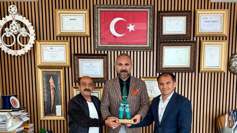 Belgeler ve Türk bayrağı önündeki odada üç kişi, ortadaki kişinin tuttuğu kupayla birlikte duruyor.