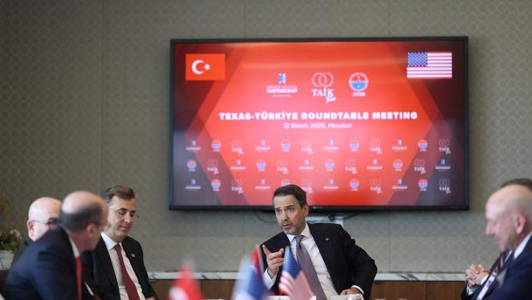 Masada oturan bir kişi elinde mikrofonla konuşurken, çevresindeki kişiler ve Türk ile ABD bayrakları görülüyor.