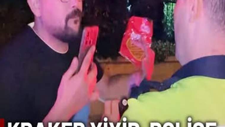 Gece çekiminde sakallı bir kişi elinde telefon ve kırmızı paket tutarken, karşısında reflektif yelekli biri duruyor.