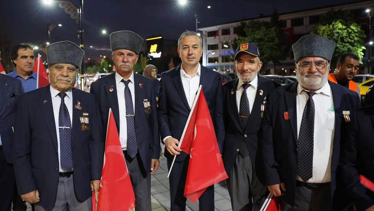 Türk bayrağı tutan, madalyalı yaşlı erkekler ve bir kişi, sokakta bir araya gelmiş.