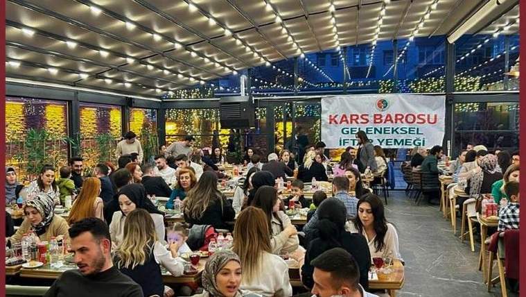 Kapalı salonda masalarda yemek yiyen kalabalık ve arkada “Kars Barosu Geleneksel İftar Yemeği” yazılı pankart.