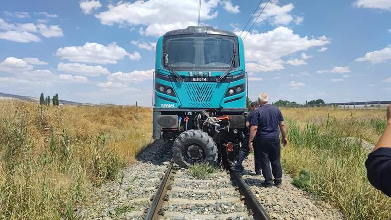 Turkuaz renkli bir raylı araç, kırsal alanda demiryolu üzerinde duruyor; yanında bir kişi ayakta bekliyor.