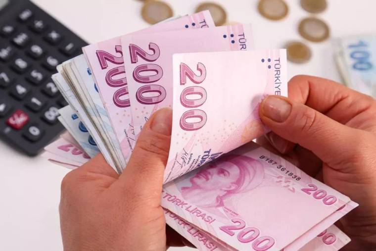 Bir kişinin elleri, 200 TL yazılı Türk lirası banknotlarını sayarken yanında hesap makinesi ve madeni paralar bulunuyor.