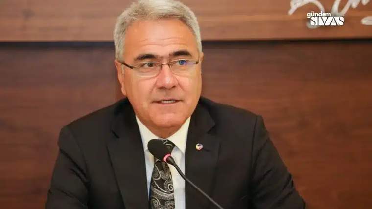 Kürsüde konuşan bir kişi, mikrofonun önünde oturuyor ve arka planda ahşap bir duvar var.
