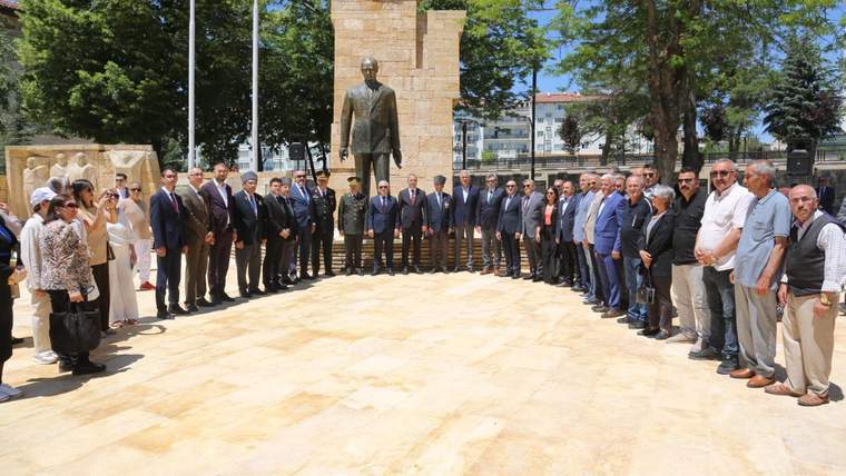 Taş sütunlu anıtın önündeki bronz heykelin çevresinde, açık alanda kalabalık bir grup insan duruyor.