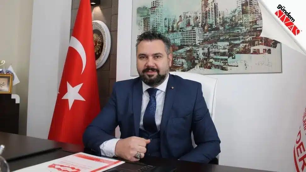 Bir masa başında oturan, takım elbise giymiş bir kişi, arkasında Türk bayrağı ve şehir manzarası olan bir tablo var.
