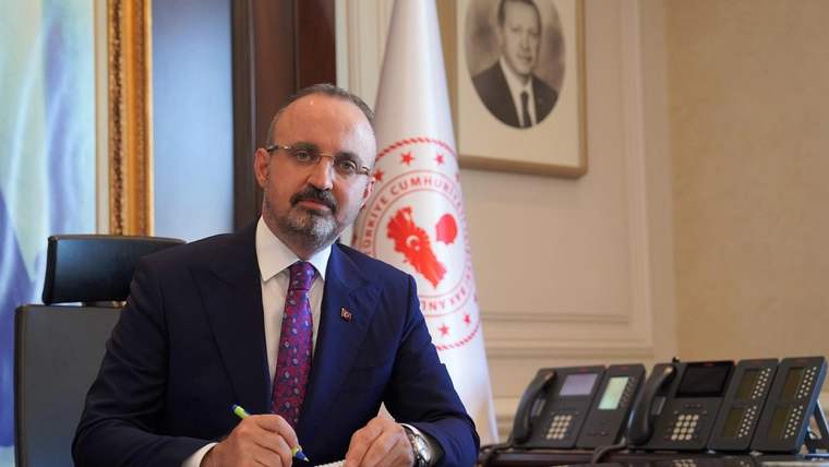 Bir masa başında oturan bir kişi, elinde kalemle not alıyor. Arkada Türk bayrağı ve bir portre var.