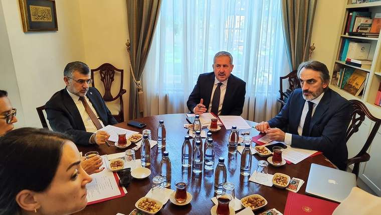 Pencereli bir odada beş kişinin masa çevresinde çay, su şişeleri ve belgelerle oturduğu toplantı