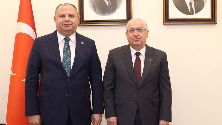 Türk bayrağı ve duvarda asılı portrelerin önünde takım elbiseli iki kişi yan yana duruyor.