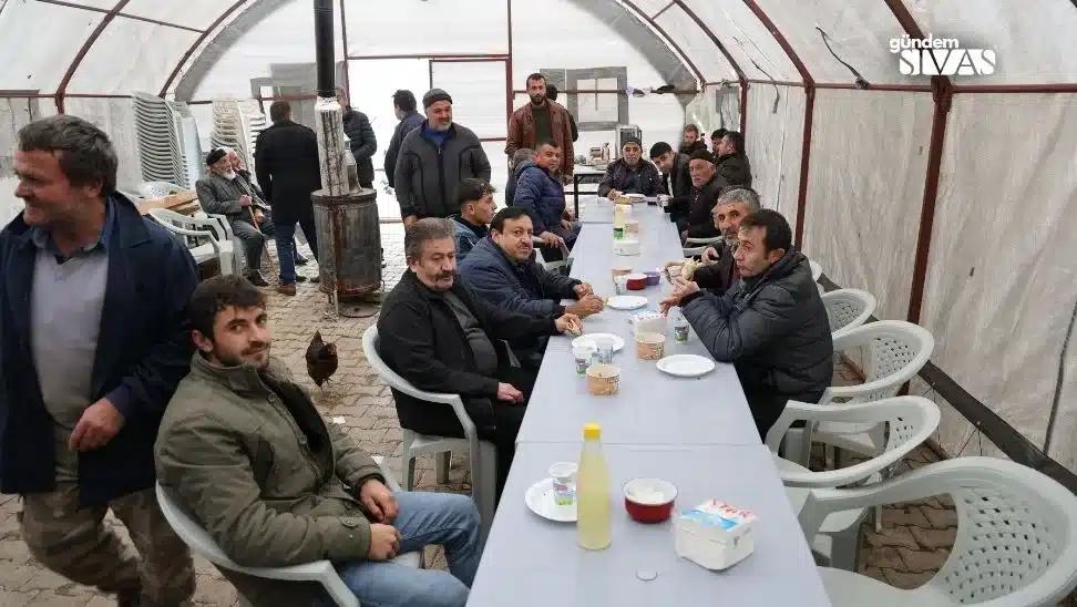 Bir grup erkek, uzun bir masanın etrafında oturmuş, yemek yiyor ve sohbet ediyor.