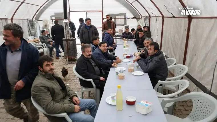 Bir grup erkek, uzun bir masanın etrafında oturmuş, yemek yiyor ve sohbet ediyor.