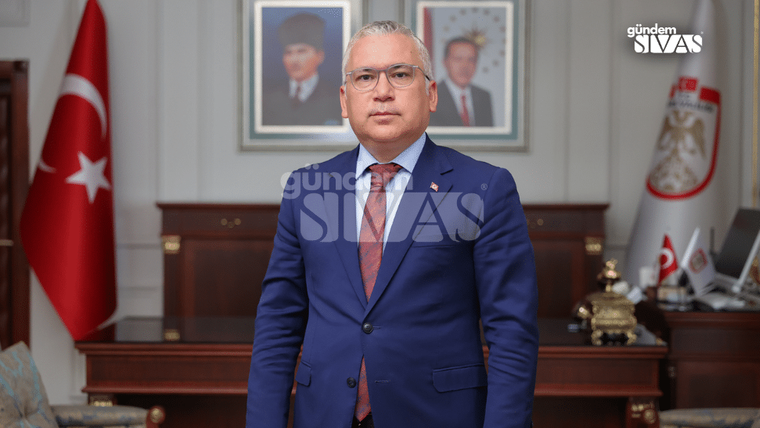 Takım elbiseli bir kişi, Türk bayrağı ve duvarda asılı portrelerin bulunduğu bir odada duruyor.