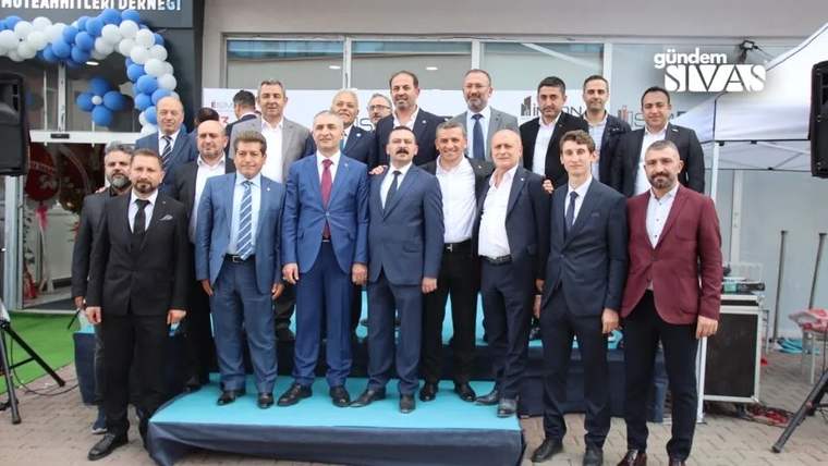 Açılış süslemeleri önünde takım elbiseli bir grup erkek, açık havada platform üzerinde birlikte duruyor.