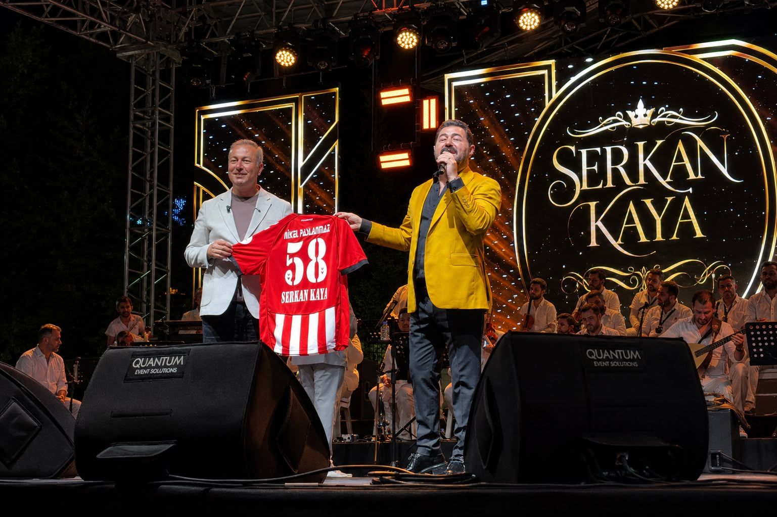 Sahnede mikrofonla konuşan bir kişi ve yanında kırmızı forma tutan bir kişi, arkada “Serkan Kaya” yazılı pano önünde.