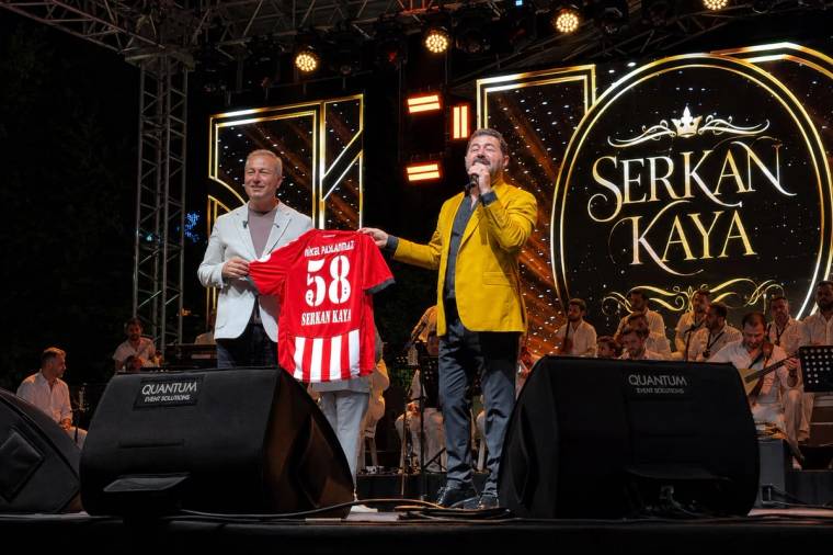 Sahnede mikrofonla konuşan bir kişi ve yanında kırmızı forma tutan bir kişi, arkada “Serkan Kaya” yazılı pano önünde.