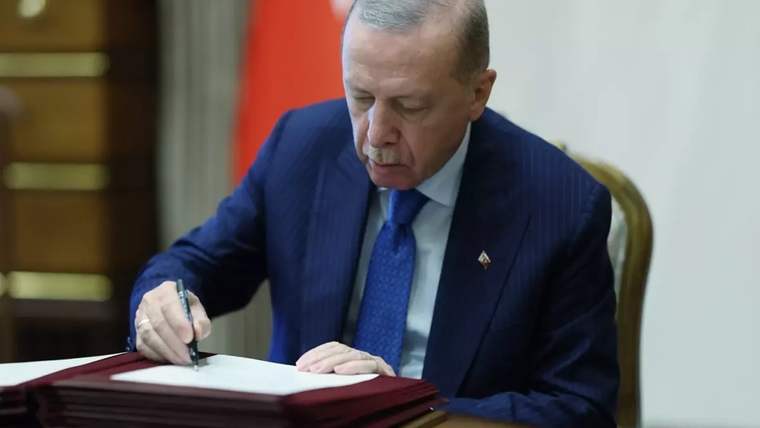 Takım elbiseli bir kişi, masa üzerindeki kalın dosyada bulunan belgeyi kalemle imzalıyor; arka planda Türk bayrağı görülüyor.