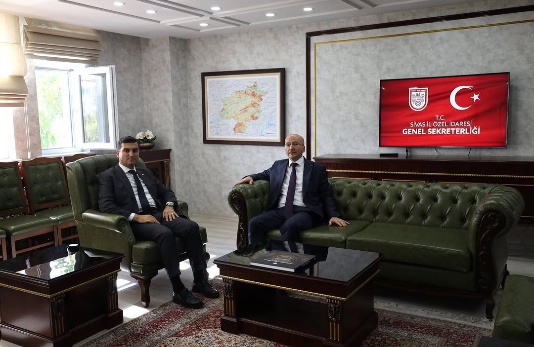 İki kişi, yeşil deri koltuklarda oturuyor; arka planda Türkiye haritası ve ekran var.