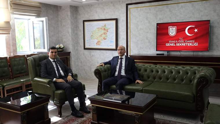 İki kişi, yeşil deri koltuklarda oturuyor; arka planda Türkiye haritası ve ekran var.