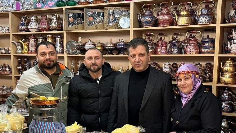 Dört kişi, arka planda çeşitli çaydanlıklar ve mutfak eşyalarıyla birlikte poz veriyor.