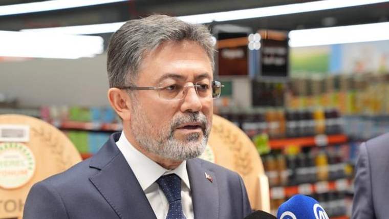 Takım elbiseli, gözlüklü ve sakallı bir kişi, market rafları önünde mikrofonlara konuşuyor.