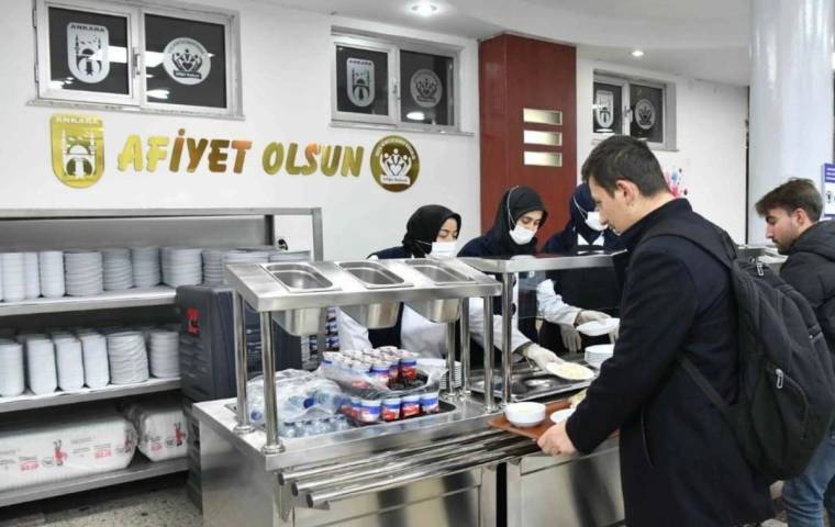 Yemek dağıtım noktasında maskeli görevliler, tezgâhtan tabak alan kişilere servis yapıyor.